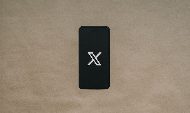 Uma imagem de um logotipo X branco exibido em um smartphone preto colocado em uma superfície marrom.