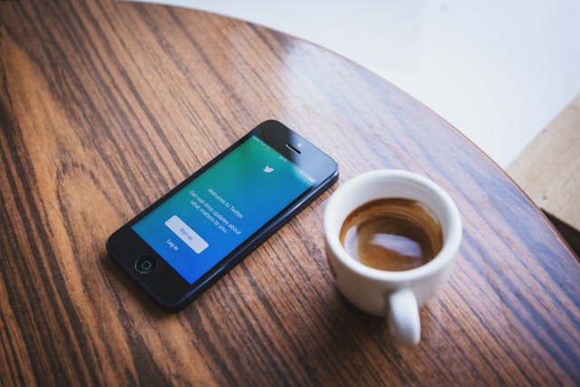 Ein schwarzes Telefon auf einem Tisch mit einer "Willkommen bei Twitter"-Seite neben einer weißen Keramik-Tasse.