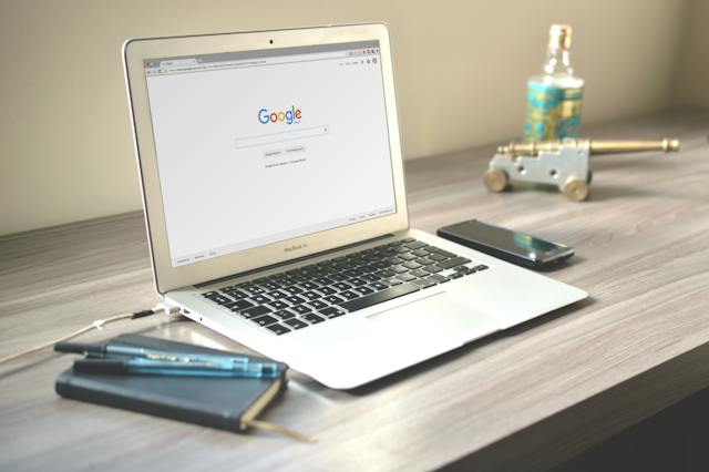 Macbook Air showing the Google Search page sitting on a grey wooden desk surrounded by pens, a notebook, and a mobile device.